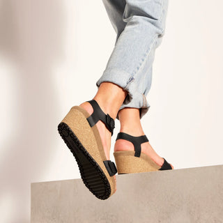 Black wedge sandals with cork soles worn by a person in light blue jeans.