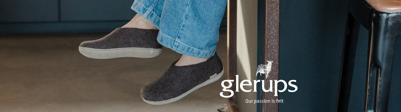 A person wearing dark gray felt slip-on shoes with light beige soles, paired with blue jeans, sitting in a modern interior setting. The brand name 'glerups' is visible in the lower right corner.
