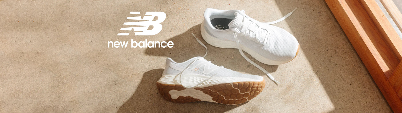 A pair of white New Balance sneakers displayed on a beige carpet with a warm wooden frame visible on the right side. The New Balance logo is printed on the left side of the image.