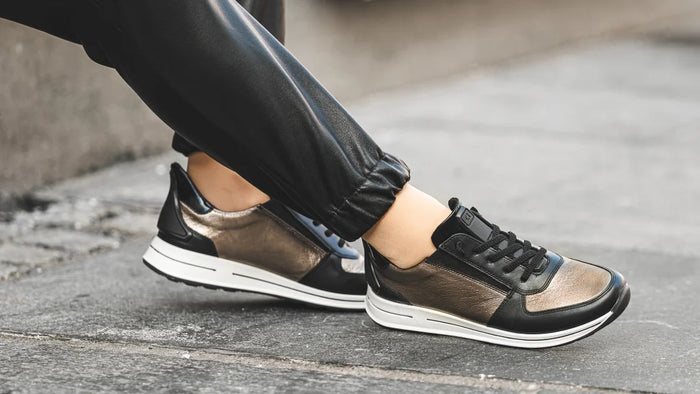 A close-up of a person wearing black leather and metallic bronze low-top sneakers with white soles, standing on a gray paved street.